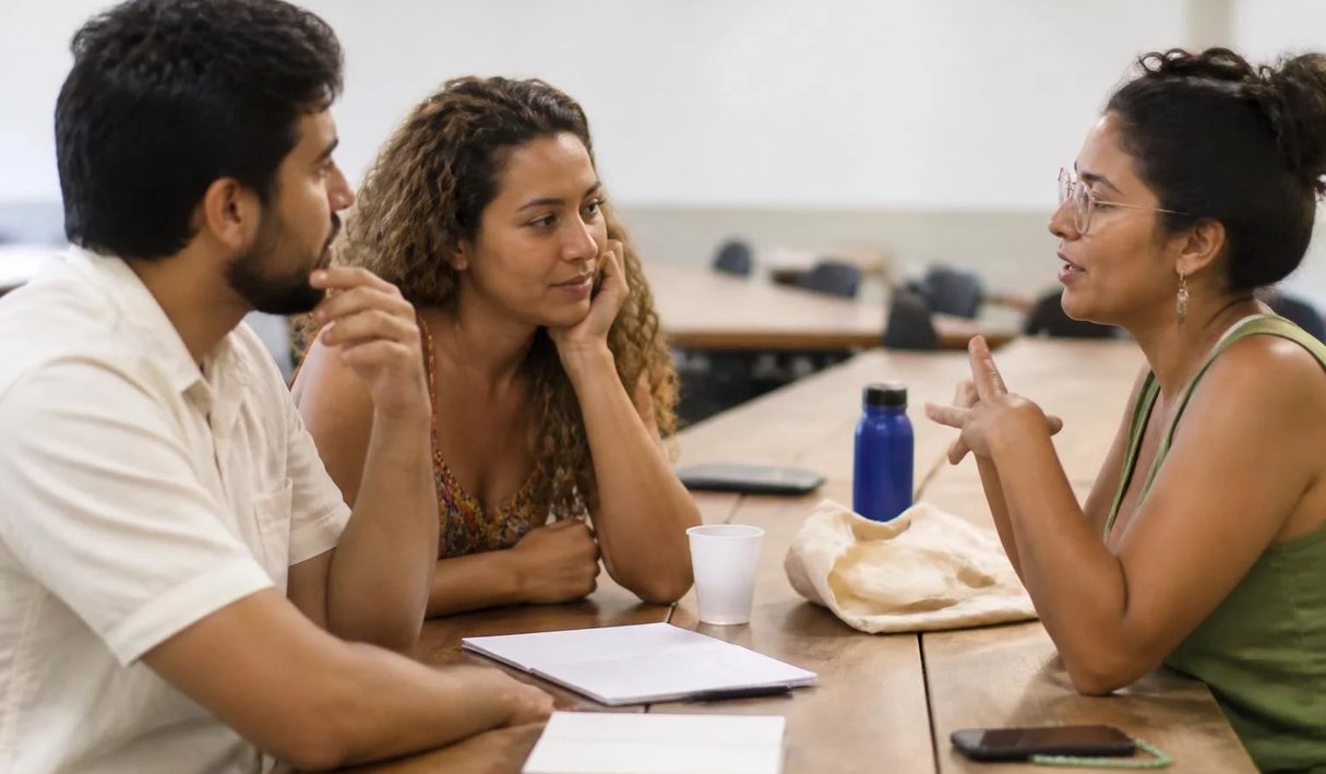 Três participantes conversam ao redor de uma mesa comprida numa sala clara de janelas altas